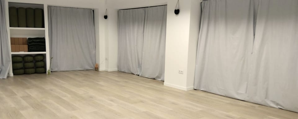 Vista amplia de la sala vacía, con suelo de madera clara, cortinas grises y la estantería de material a la izquierda.