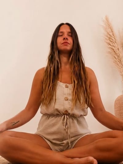 Natalia sentada con las piernas cruzadas y los ojos cerrados, meditando en una sala de paredes claras.